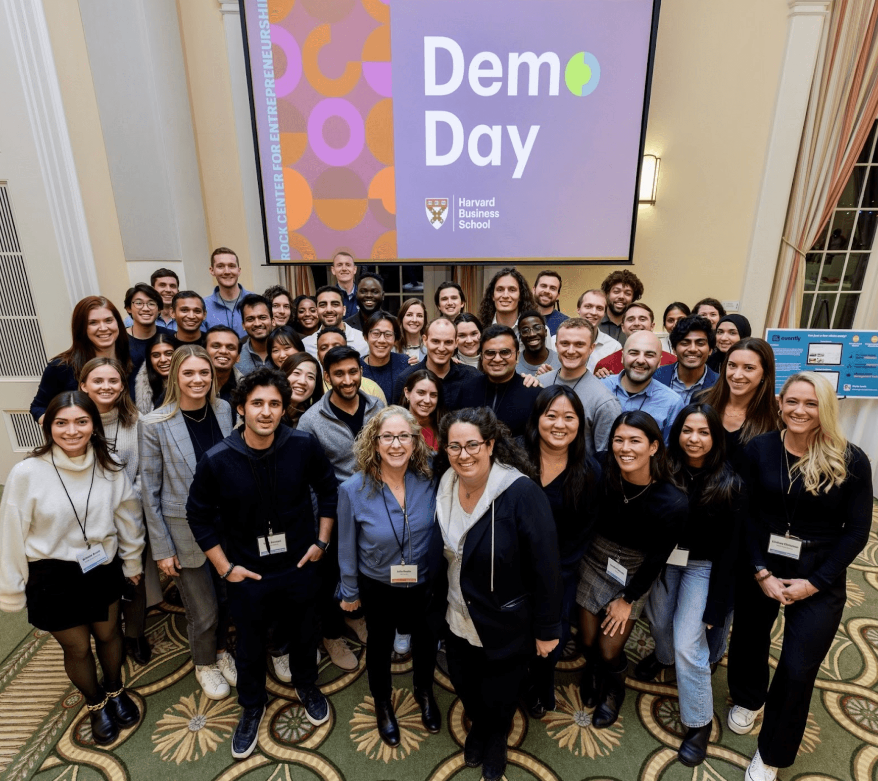 Group photo of attendees at Harvard Business School's Demo Day, smiling in front of a screen displaying 'Demo Day' and the HBS logo.